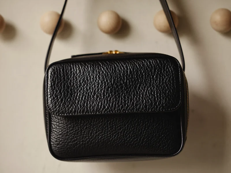 Tracolla nera goffrata con zip in ottone appesa a un semplice piolo di legno.