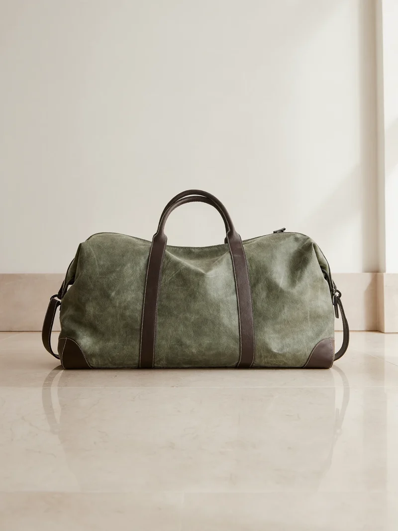 Duffel in tela cerata verde oliva con manici in pelle, ripresa dal basso su pietra chiara.